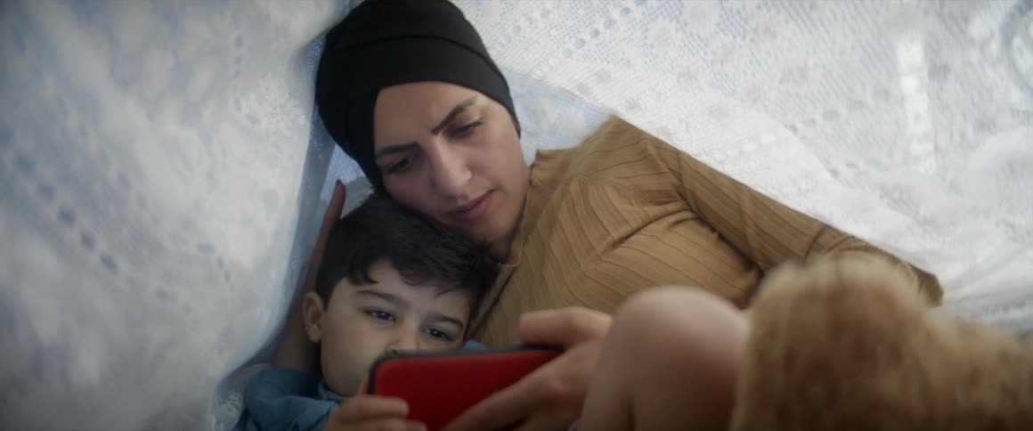 A woman and her son watch a video on a smartphone while lying under a bed sheet