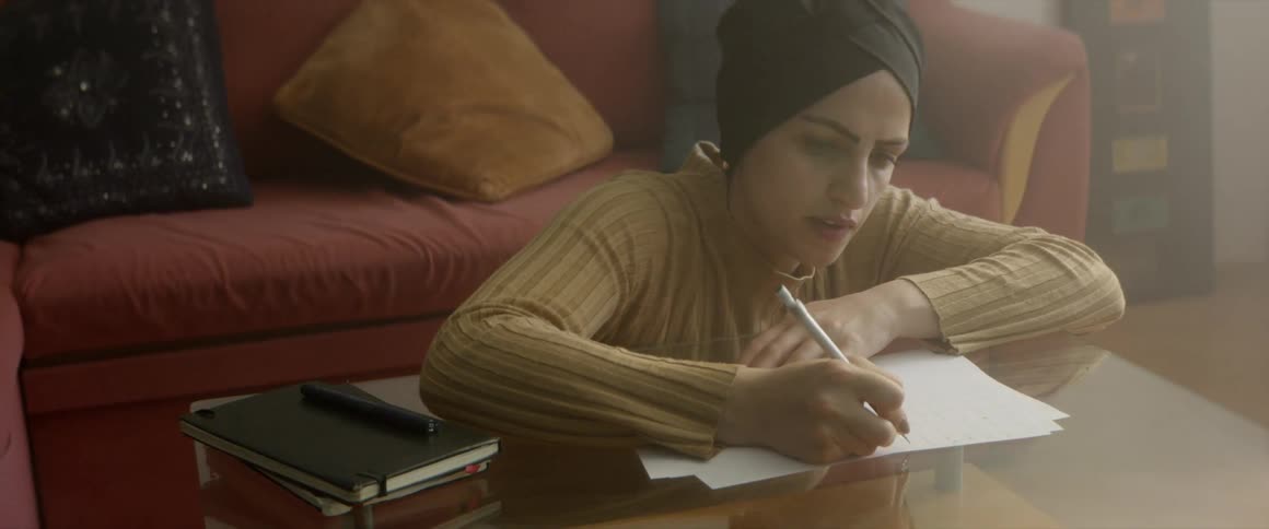 Woman wearing a long sleeved shirt and head covering sits on the floor at a coffee table writing on a piece of paper beside a book in front of a couch with throw pillows