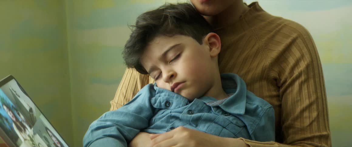 A woman holds her sleeping son in her lap as she looks at a tablet