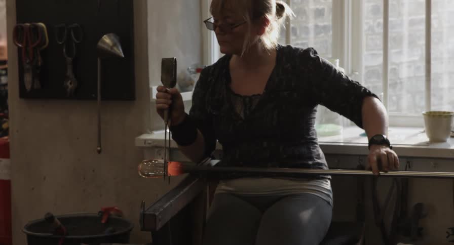 A woman shapes hot glass with tools