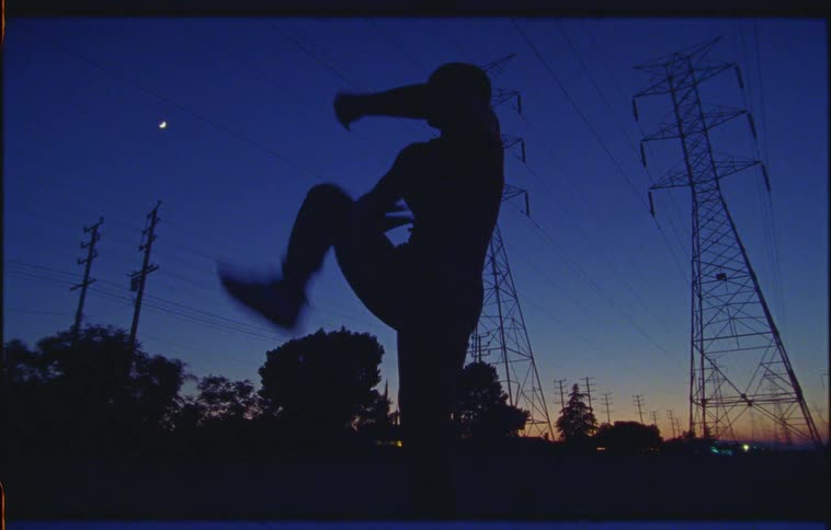 Woman dances outdoors under electric transmission towers with afterglow and town lights on horizon