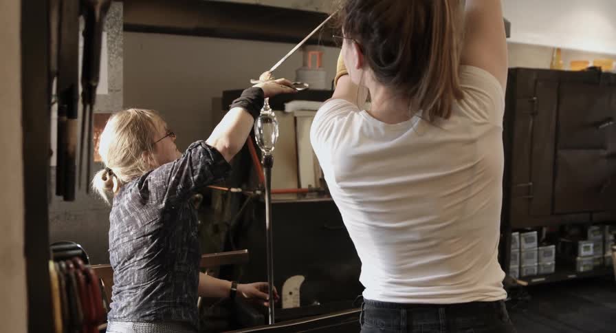 Two women work together to  put a ball of hot glass onto a glass piece being molded