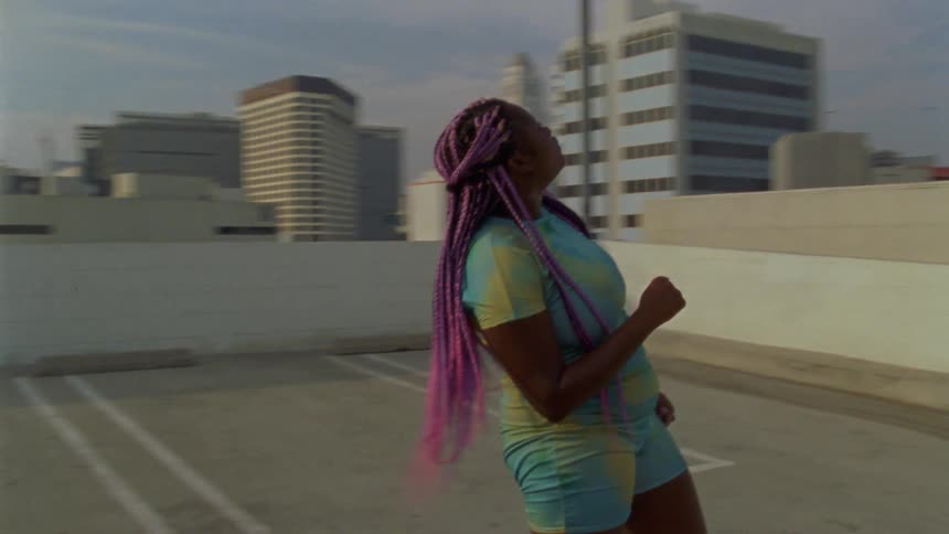 A woman with purple braids dances on top of a parking garage next tall skyscrapers