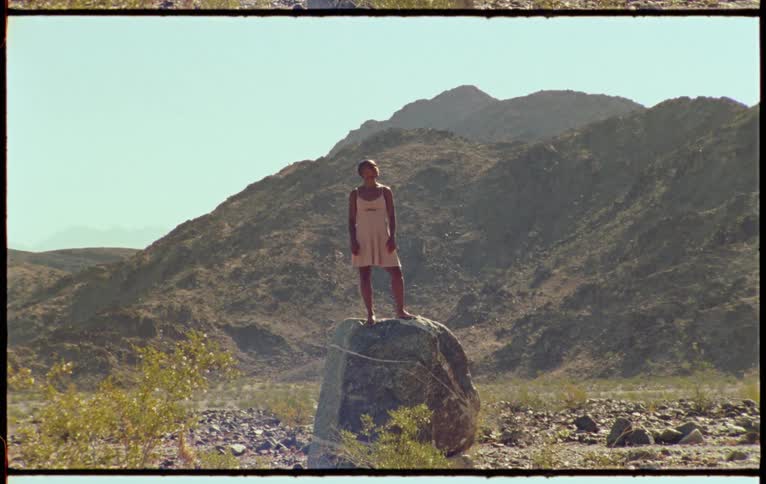 A young woman stands on a large rock and looks up at the sky