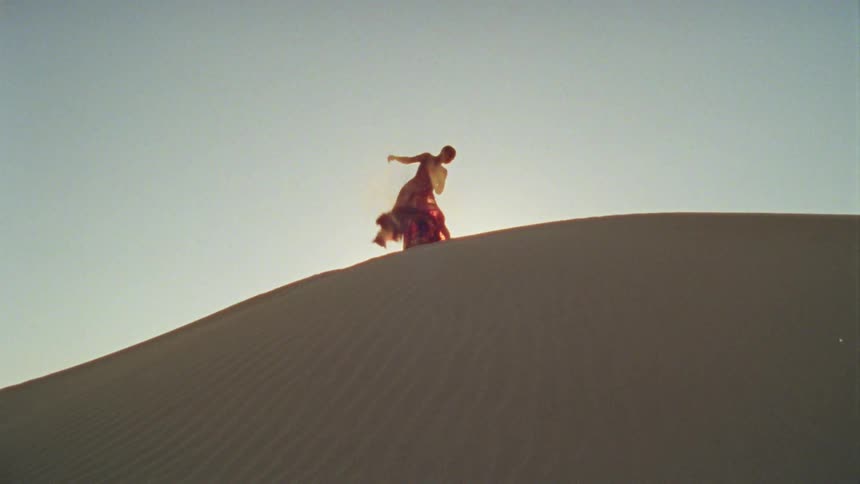 A woman in a long red dress dances atop a sand dune in the desert during the day