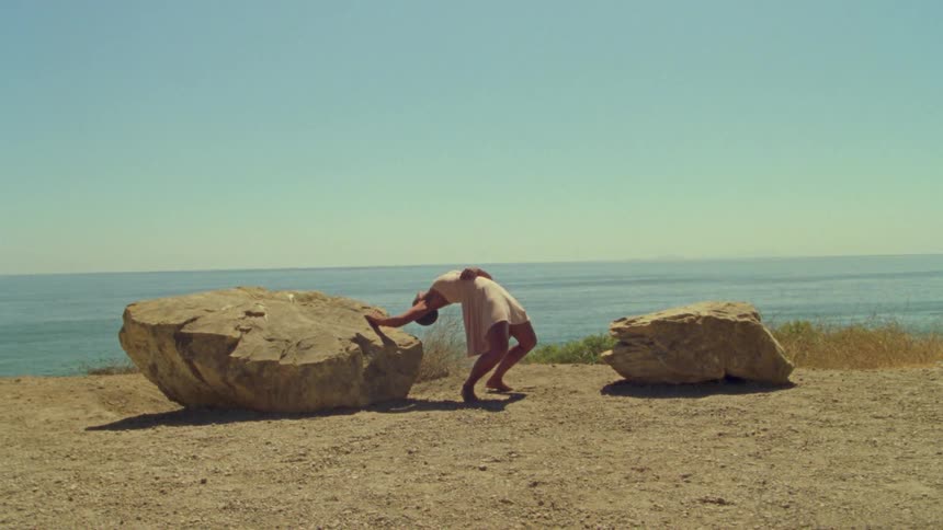A woman on the beach dancing next to rocks with the ocean sky and clouds in the background