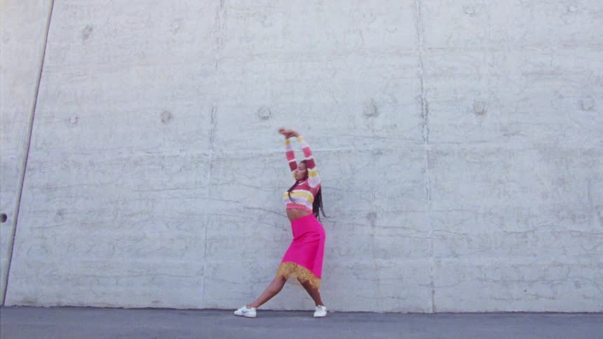 A woman in a bright pink skirt dances outside in front of a concrete wall
