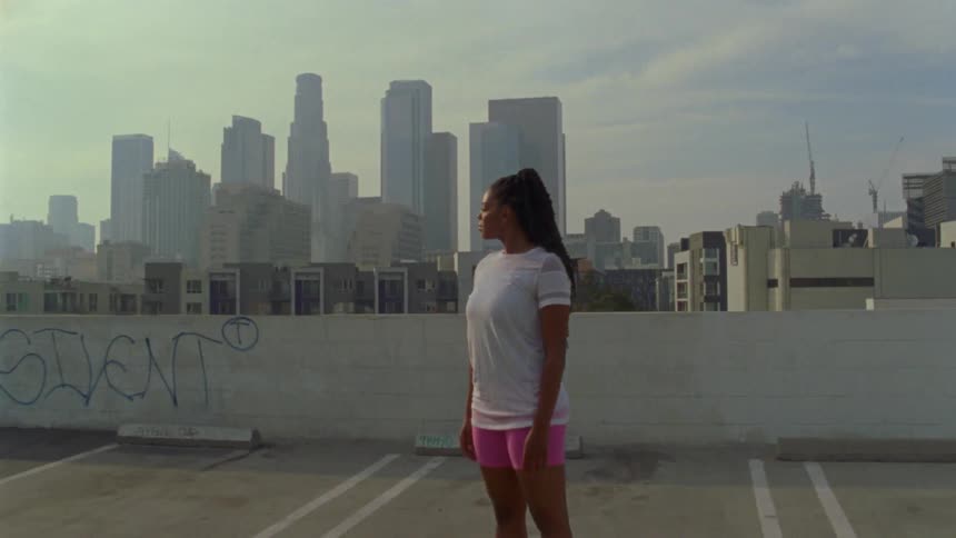 A young woman standing in a parking lot on the roof of a parking garage turns her head to look into the approaching camera