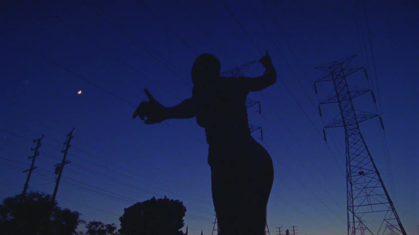 A woman dancers outdoors beneath a pair of transmission towers at sunset