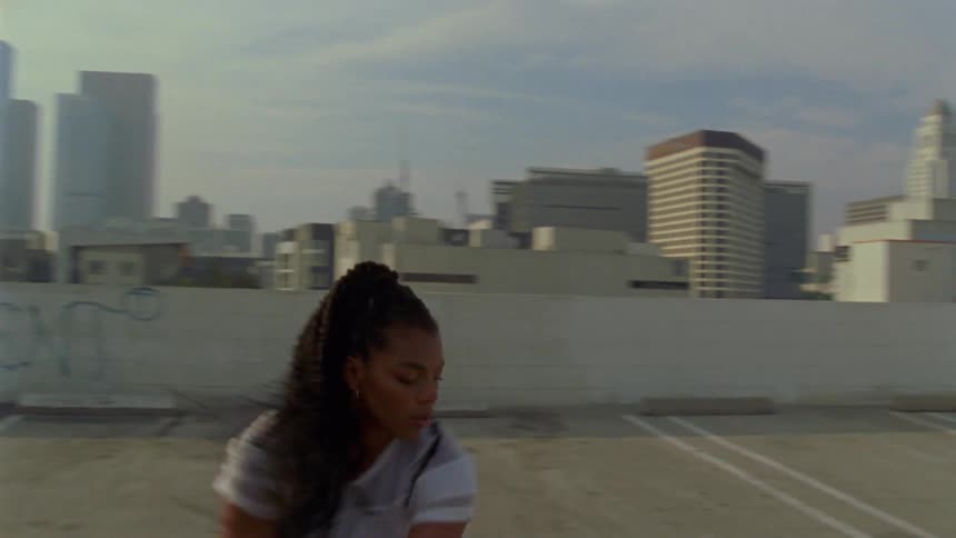 A black female dances on a rooftop in a downtown area of a city with tall buildings in the background