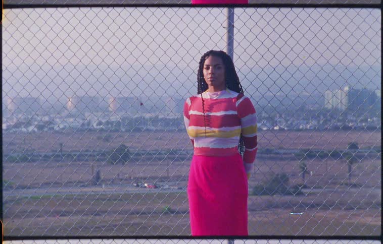 A woman with a sweater and a skirt standing by a fence with buildings the sky and clouds in the background