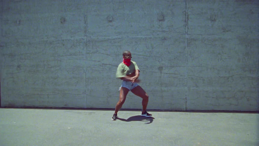 Bald woman wearing a bandana over her face dances energetically in front of a concrete wall