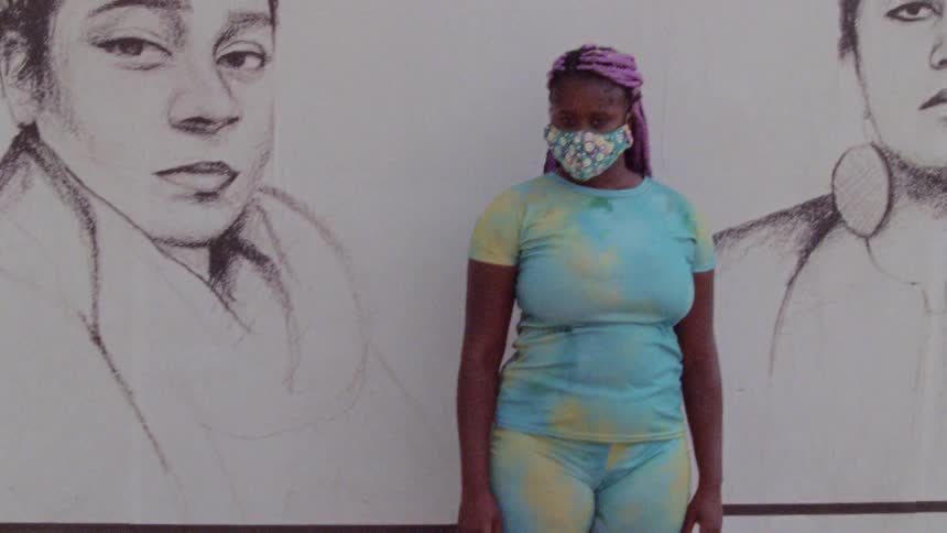 A woman wearing a face mask stands in front of a mural
