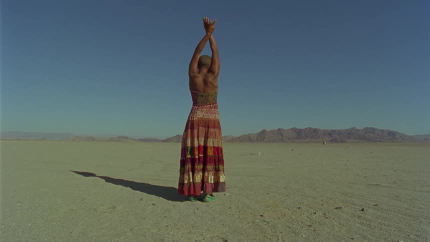 Bald woman wearing a dress stands in the desert stretching her arms up