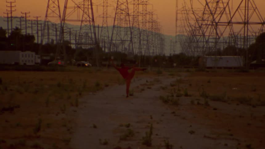 Person dances in field under electric transmission towers with mountains on horizon sitting under red tinted sky