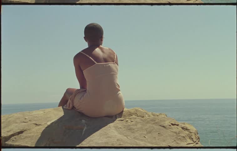 Bald woman wearing a pink dress sitting in a rock near the ocean