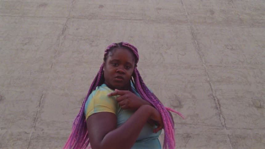 A woman performs a dance move in with her hands and stares at the camera outdoors in front of a concrete wall at daytime