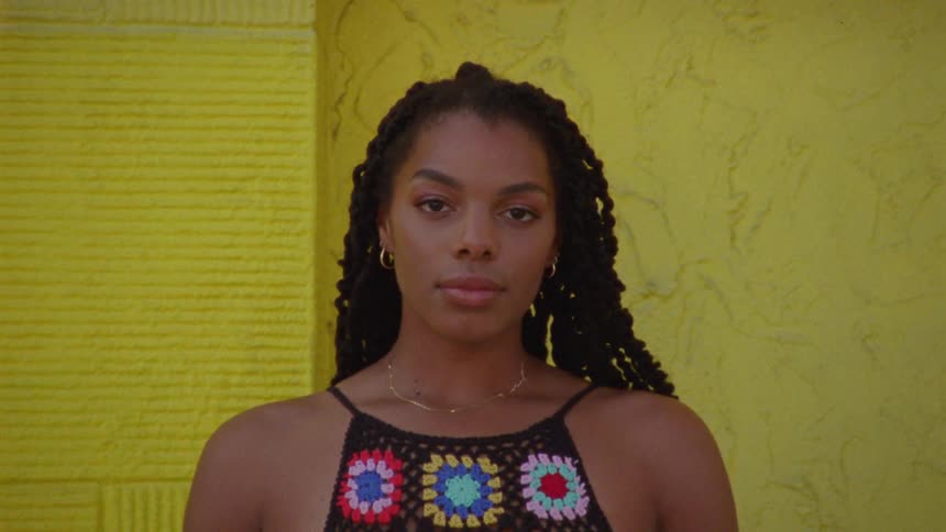 A woman out of focus flips her hair and as the camera comes into focus looks at the camera with a neutral expression
