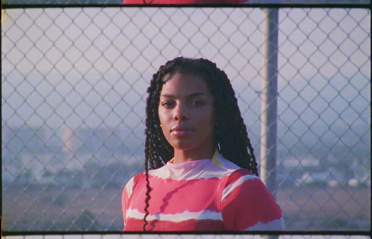 A woman with long braids wearing a red and yellow dress stands with a chain link fence behind