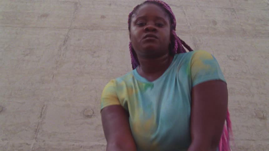 A woman approaches and kneels in front of the camera before gesturing with her hand and chin