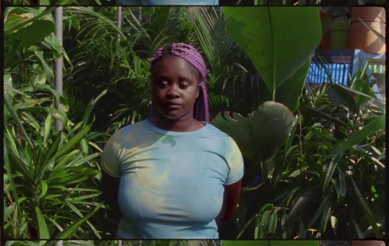 Woman with long pink hair stands in front of plants looking at camera