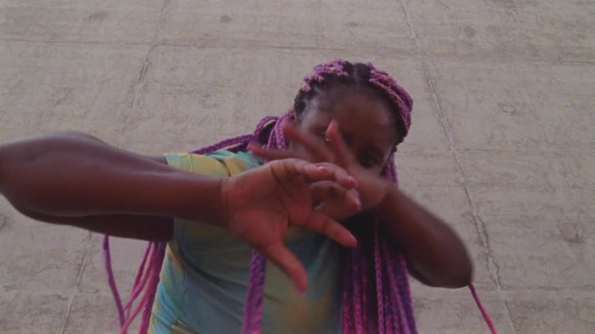 Woman with pink and purple braid extensions dances against concrete block background