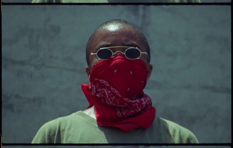 Woman with a red bandana over her face and sunglasses standing in front of a concrete wall