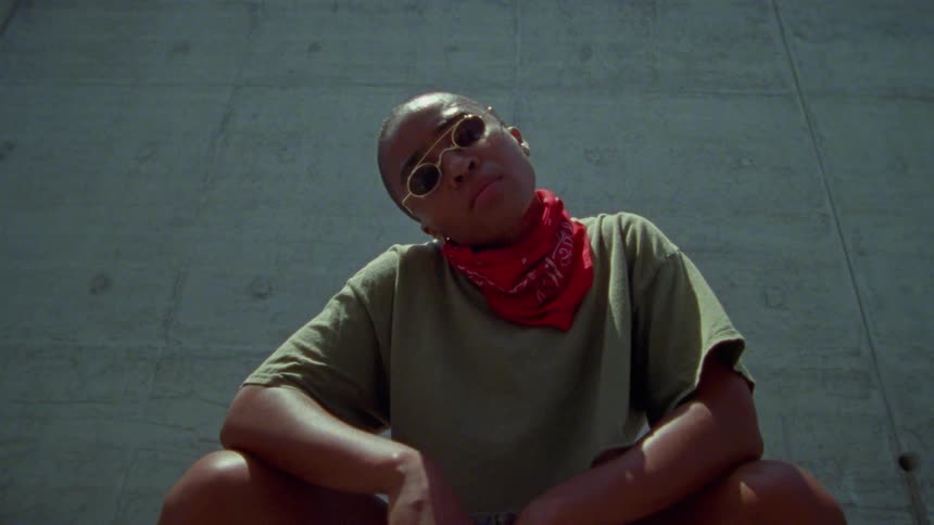 A woman with sunglasses and a bandanna walking up and stooping down with a wall in the background