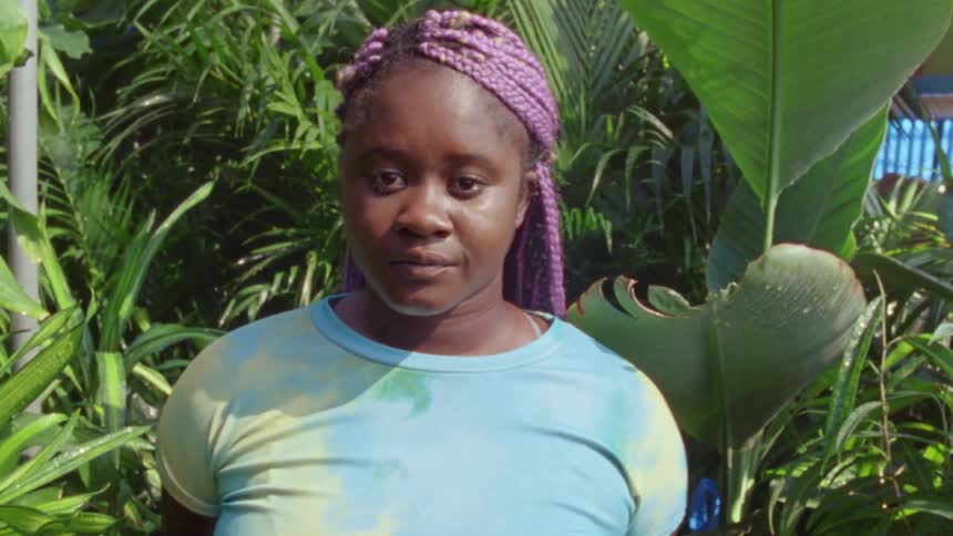 A woman looks into the camera while standing among tall vegetation outside a building during the day