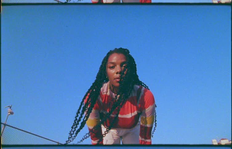 A young woman bends over to look into the camera as her hair waves in the wind under a clear blue daytime sky