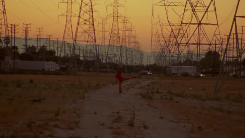 A person dances in a field in front of electrical towers