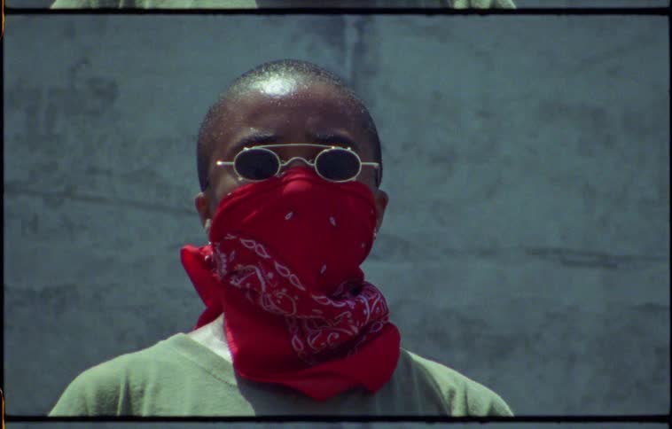 Person sweating outside wearing sunglasses and bandana over face stands next to wall
