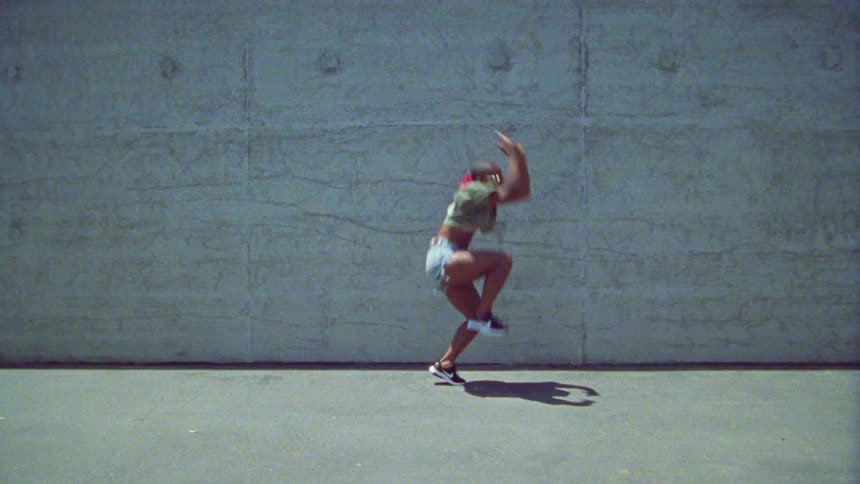 A woman performing hip hop dance moves outdoors in front of a large concrete wall at daytime