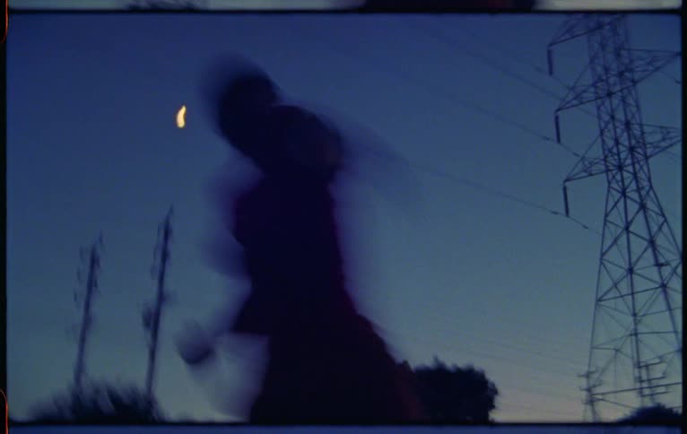 A woman dances outdoors beneath a transmission tower at sunset