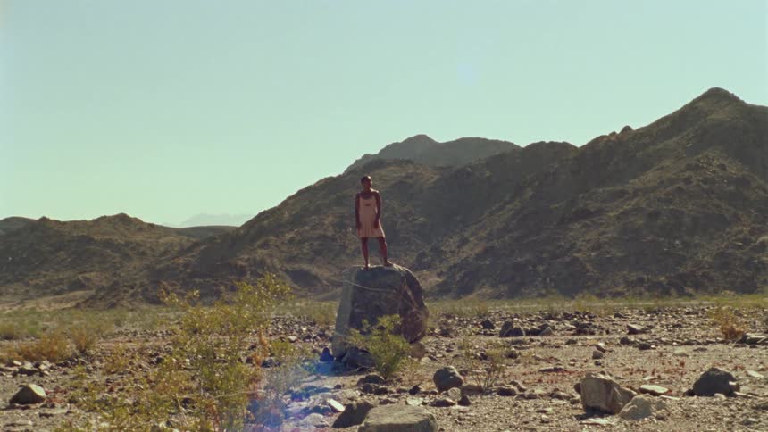 Woman wearing sundress stands on large boulder in field near rocky hill