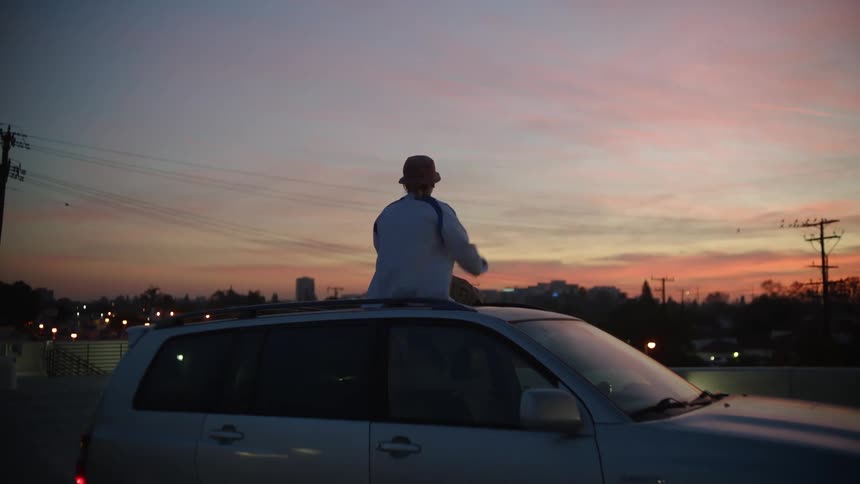 A man sits on the roof of his car and raises his middle fingers towards the city skyline