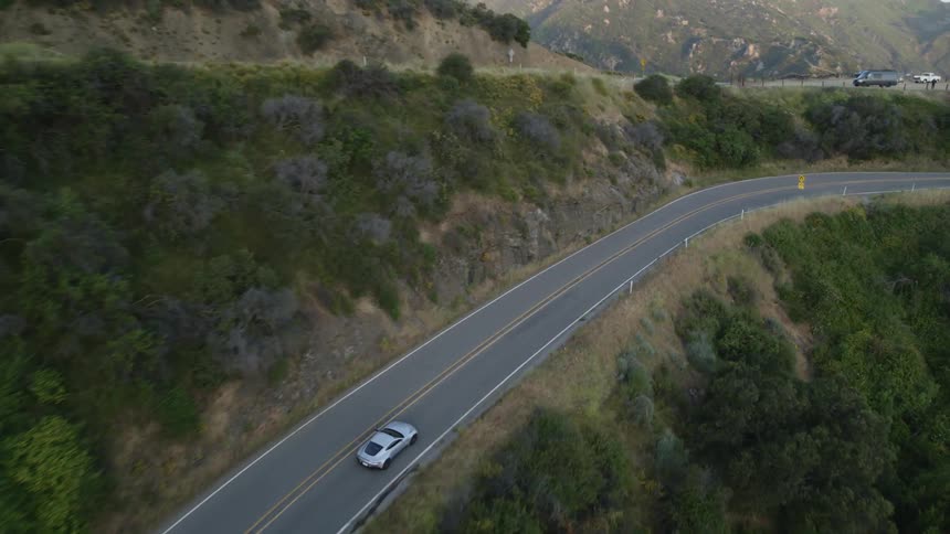 A silver sedan drives along a curvy mountain road that overlooks houses