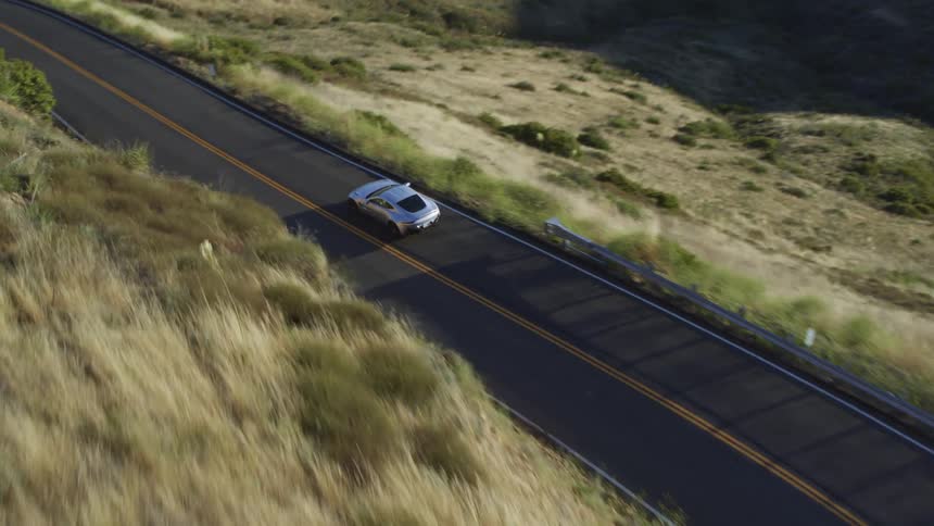 Sports car winds along road bordered by steep slope passing hills and mountains