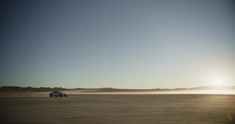 A silver sports car driving off road in a desert with a wide open view of the horizon as the sun rises