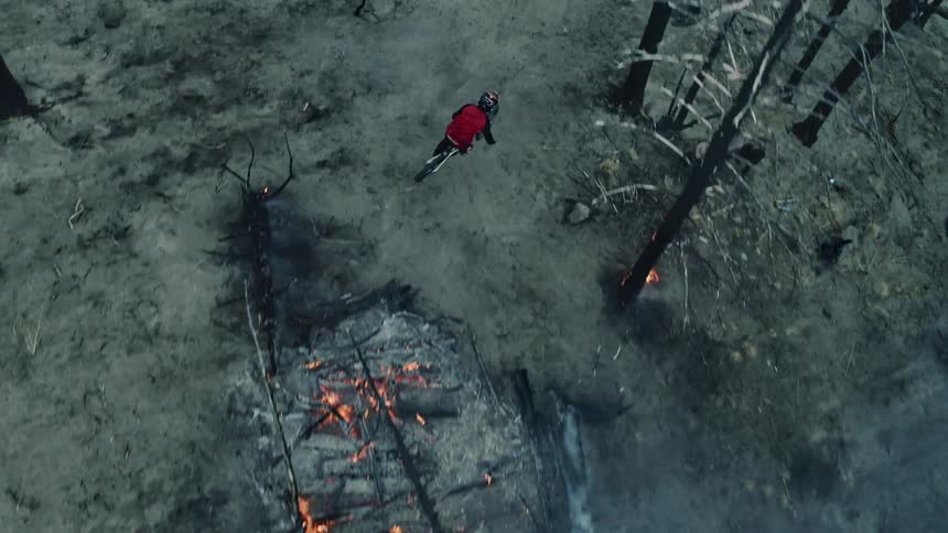 Drone angle of a mountain biker jumping over a fire in a burning forest.