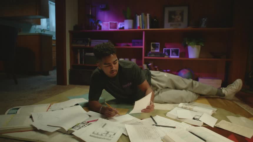A teenage boy lies on the floor at home with papers strewn about him and writes as he does homework