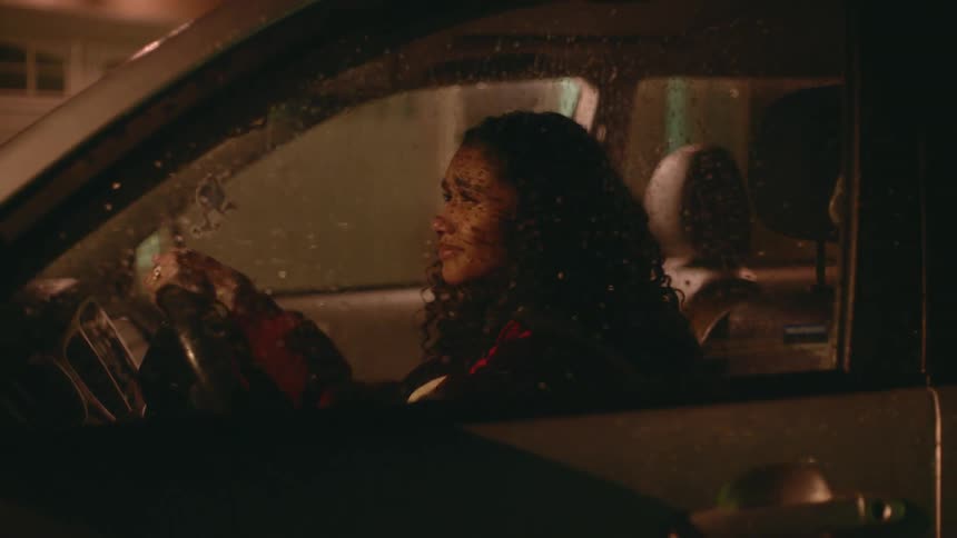 Young woman sitting in a parked car in front of a house at nighttime crying while she holds the steering wheel