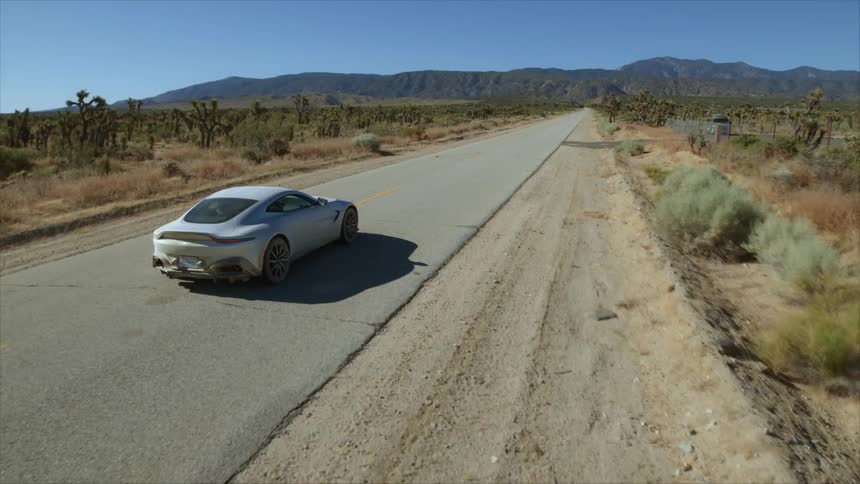 Sports car passes homestead set in scrubby field while heading toward mountains
