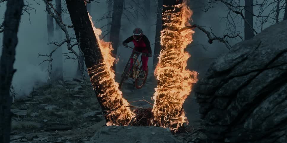 A person wearing a helmet jumps a mountain bike between burning trees in a forest