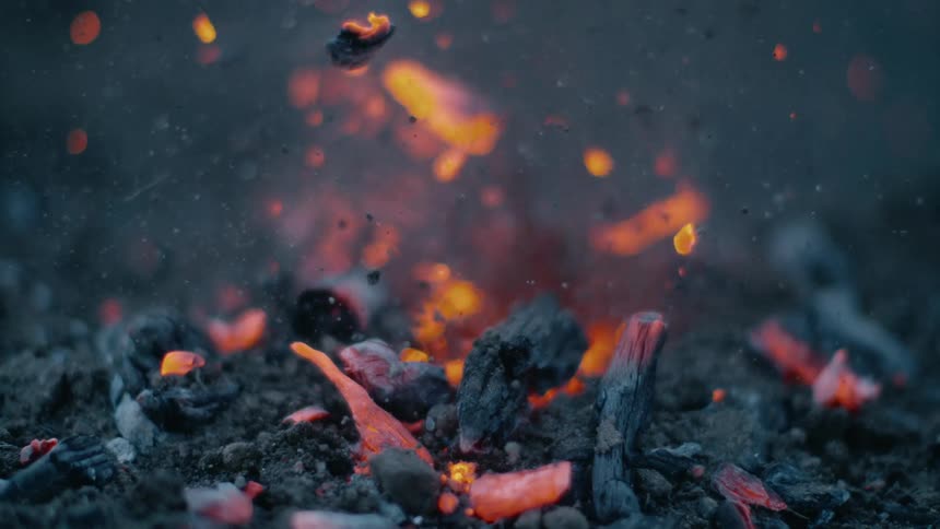 Embers and burning bits of wood fall from the air around a flame surrounding by piles of ash