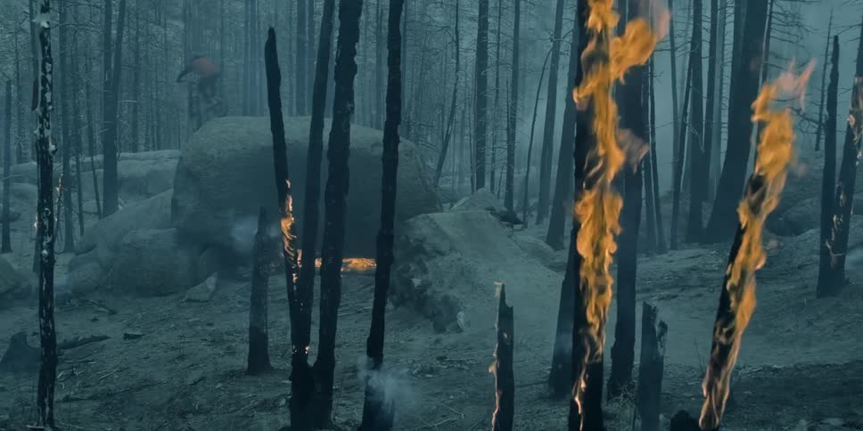 A man jumps with a mountain bike on and off a rock in a burning forest