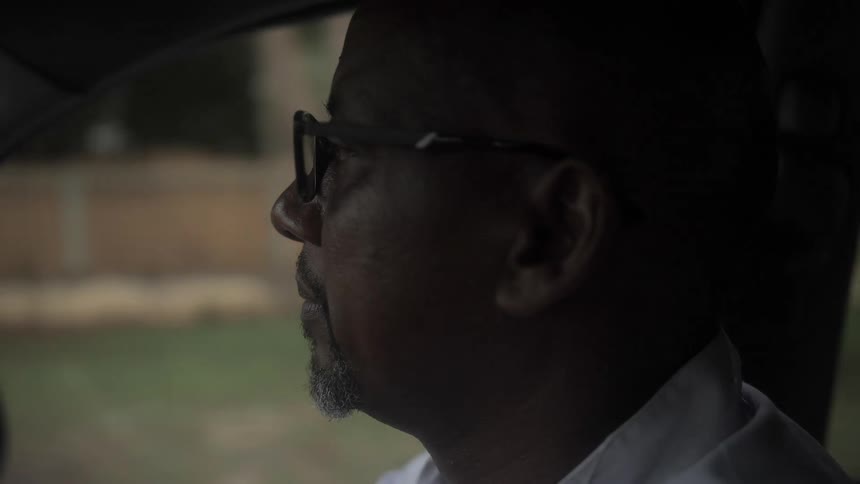 A man with glasses driving in a vehicle with a fence trees the sky and clouds in the background