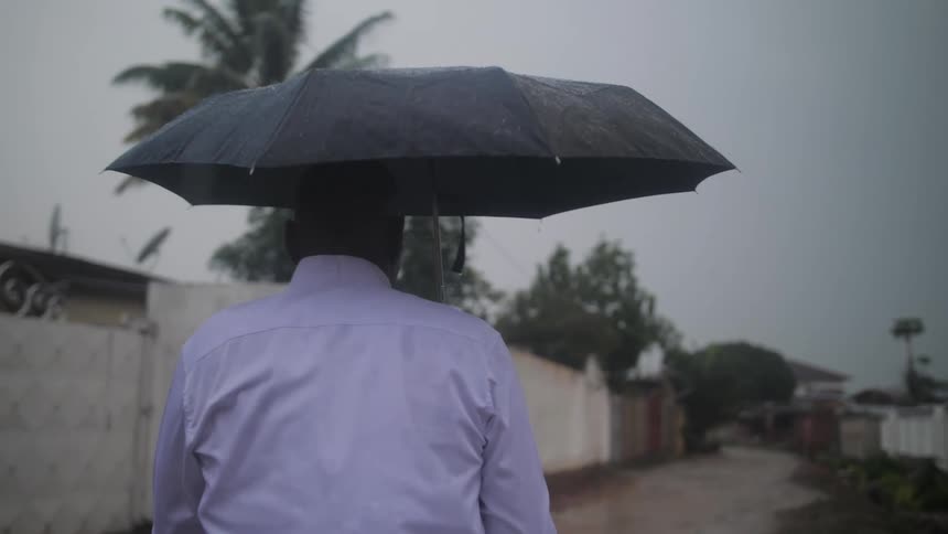 A man holds a black umbrella over his head shielding from the rain while walking down an alleyway in a neighborhood