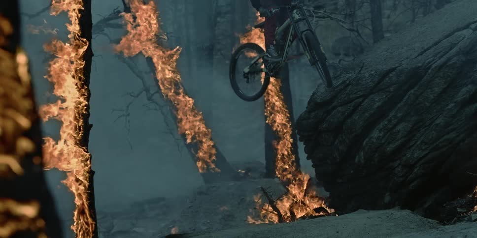 An extreme bike rider bonks front tire off rock with trees engulfed in flames.