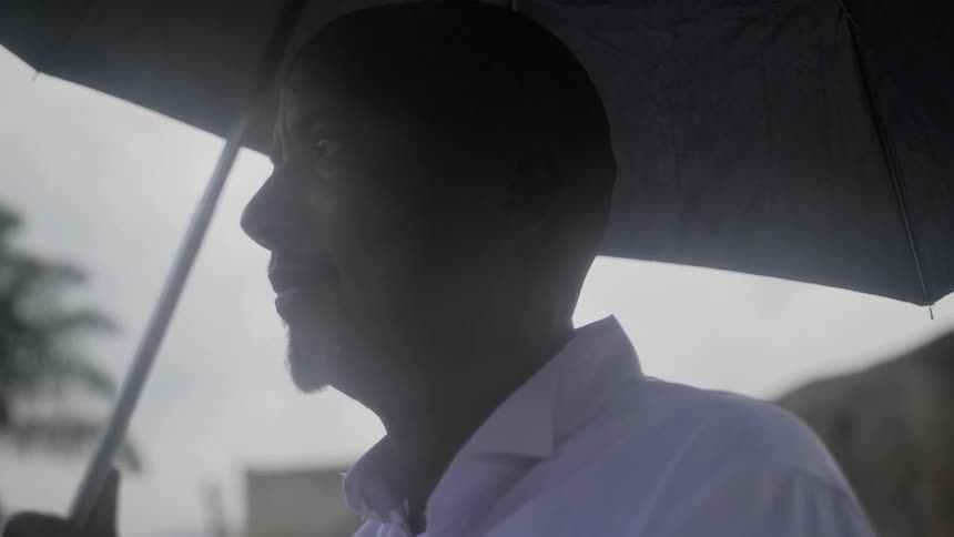 A man stands in the rain while holding an umbrella above his head and looking off into the distance
